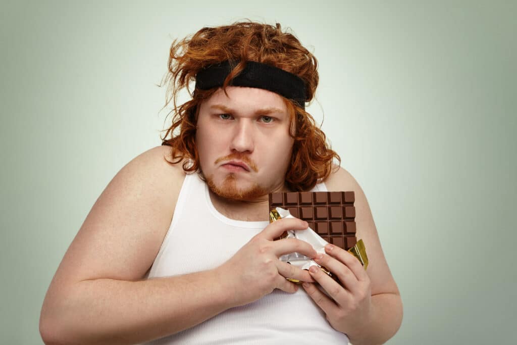 Greedy obese fat young man wearing sports band on curly ginger hair, grimacing, looking at camera with suspicious mistrustful expression, about to eat up large bar of chocolate, holding in tight
