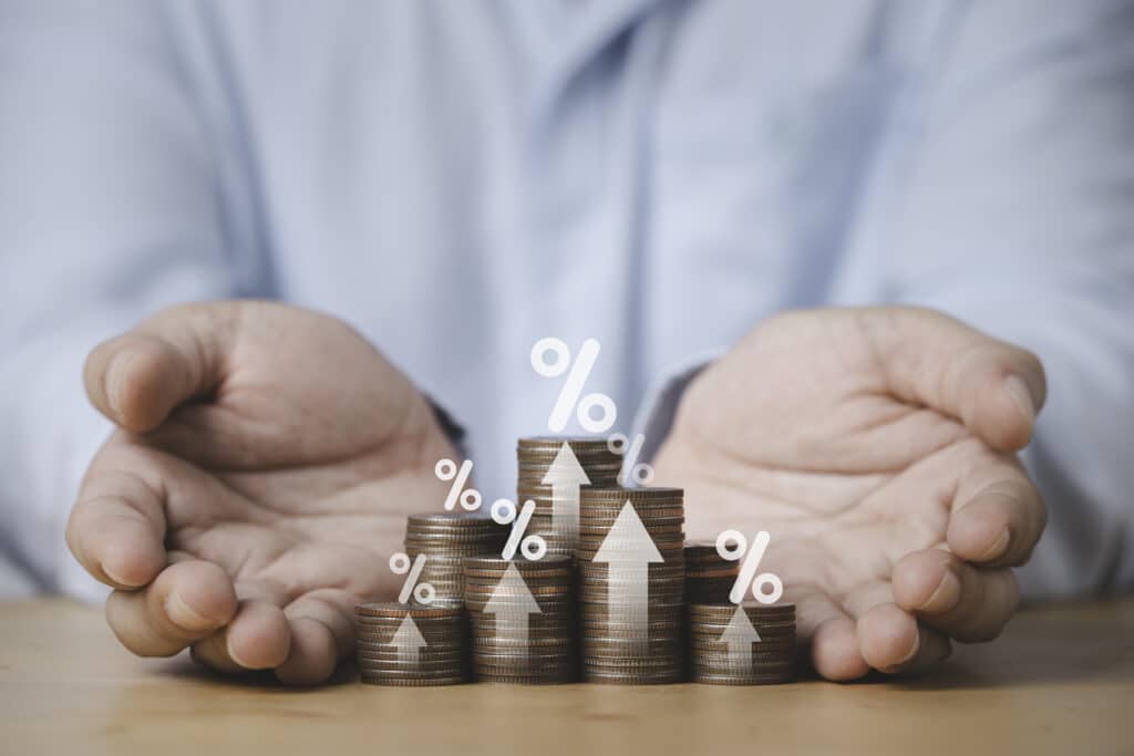 Two hand giving heap of coins money with up arrow and percentage ปัจจัยที่ส่งผลต่อดัชนีดอลลาร์สหรัฐ : อัตราดอกเบี้ย